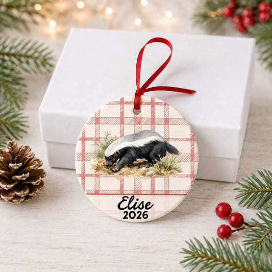 Personalized honey badger ceramic ornament with name and year on red plaid in front of a white gift box with pinecones and red berries