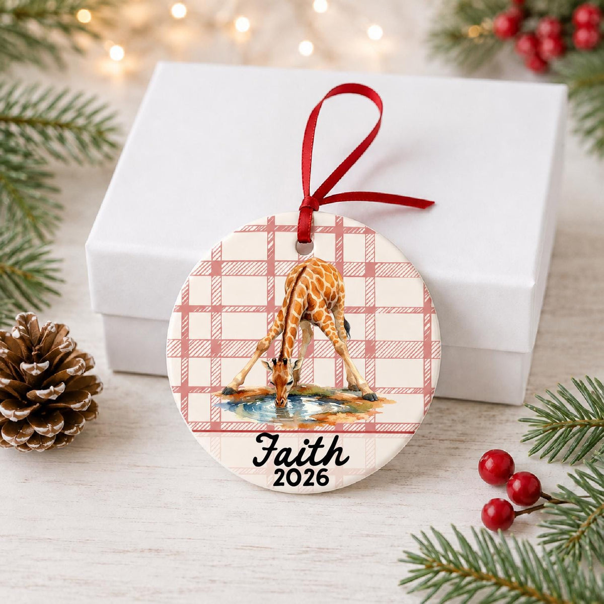 Personalized giraffe ceramic ornament with name and year on red plaid in front of a white gift box with pinecones and red berries