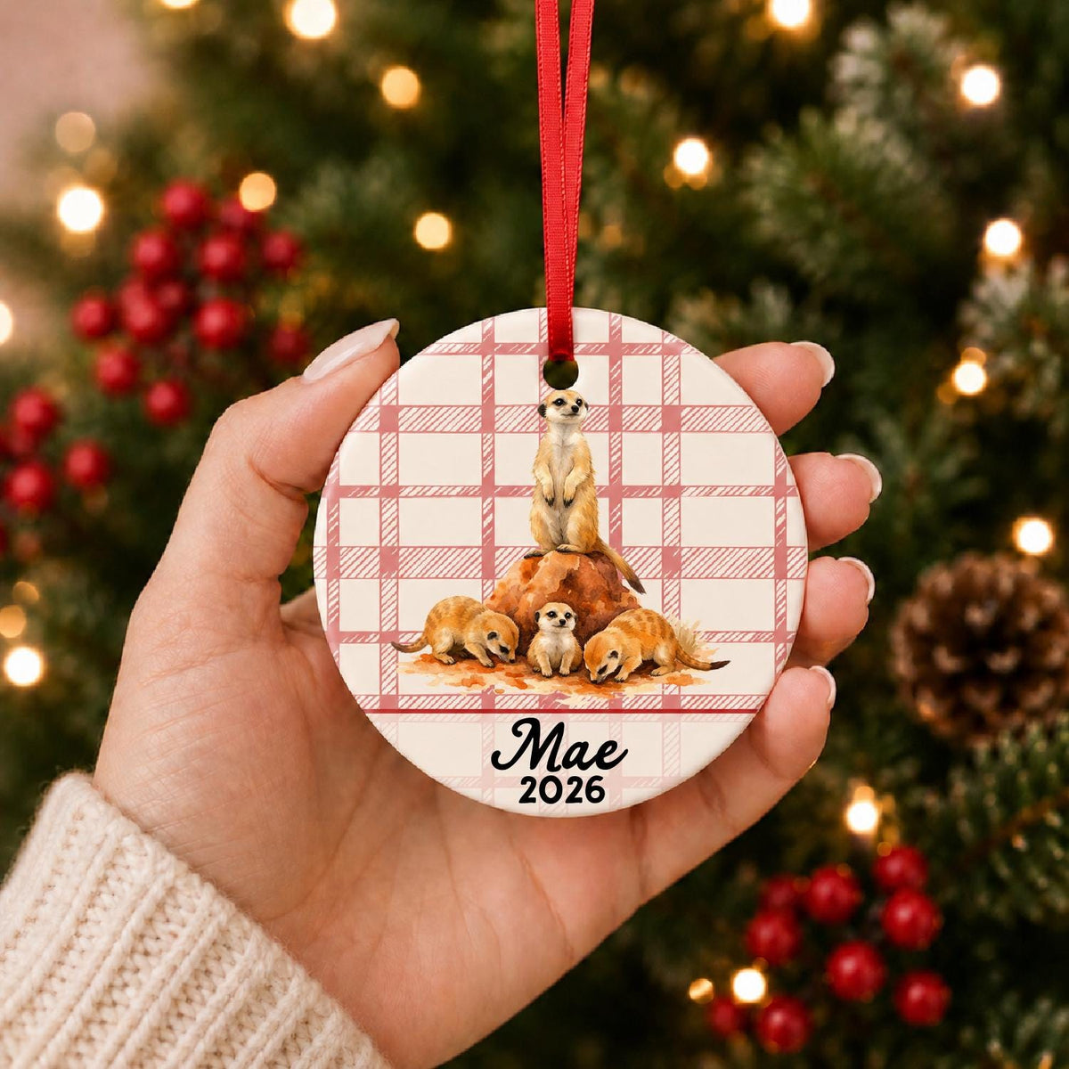 Hand holding the personalized meerkat ceramic ornament with name and year on red plaid in front of a Christmas tree