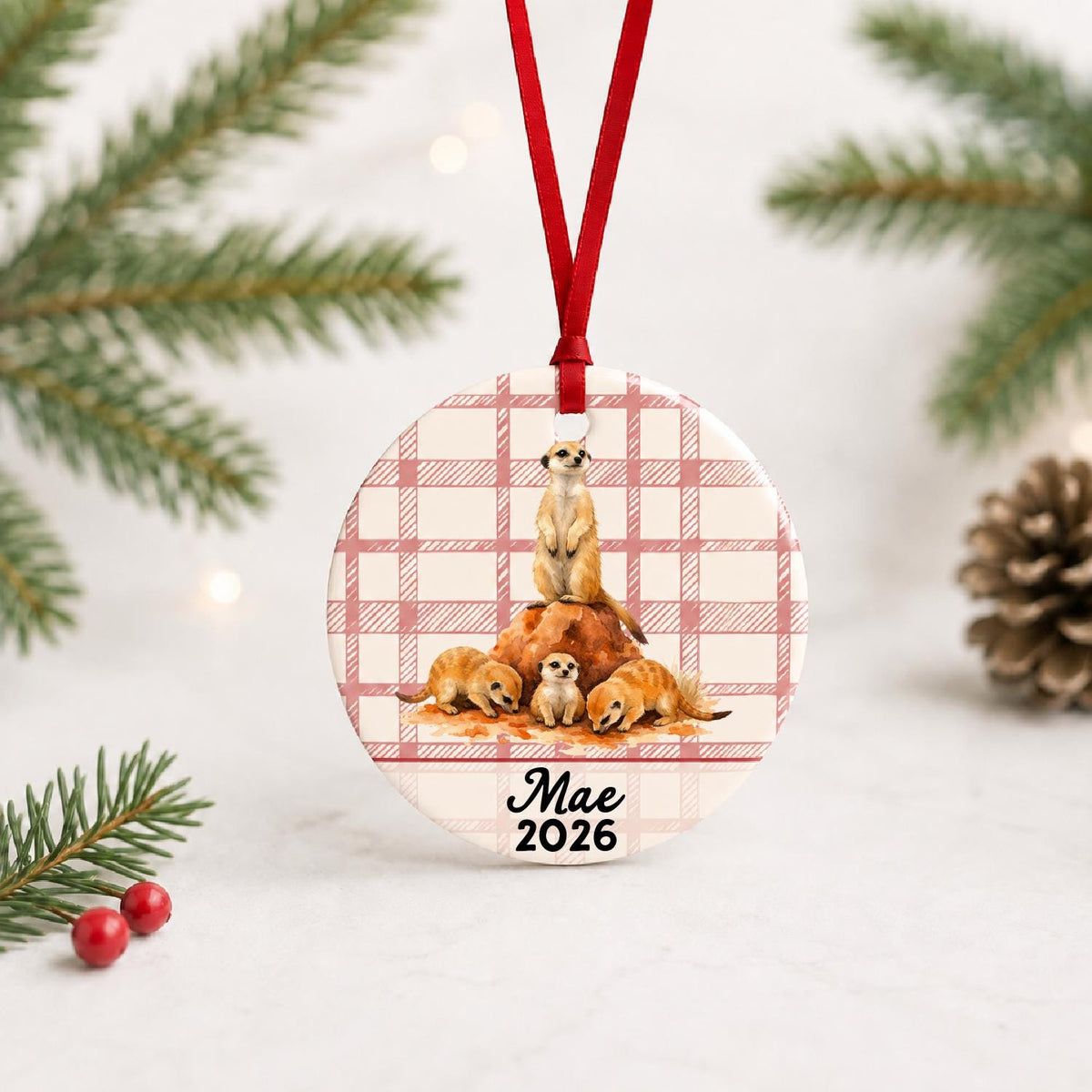 Personalized meerkat ceramic ornament with name and year on red plaid with red ribbon on a white surface with pine branches and pinecones