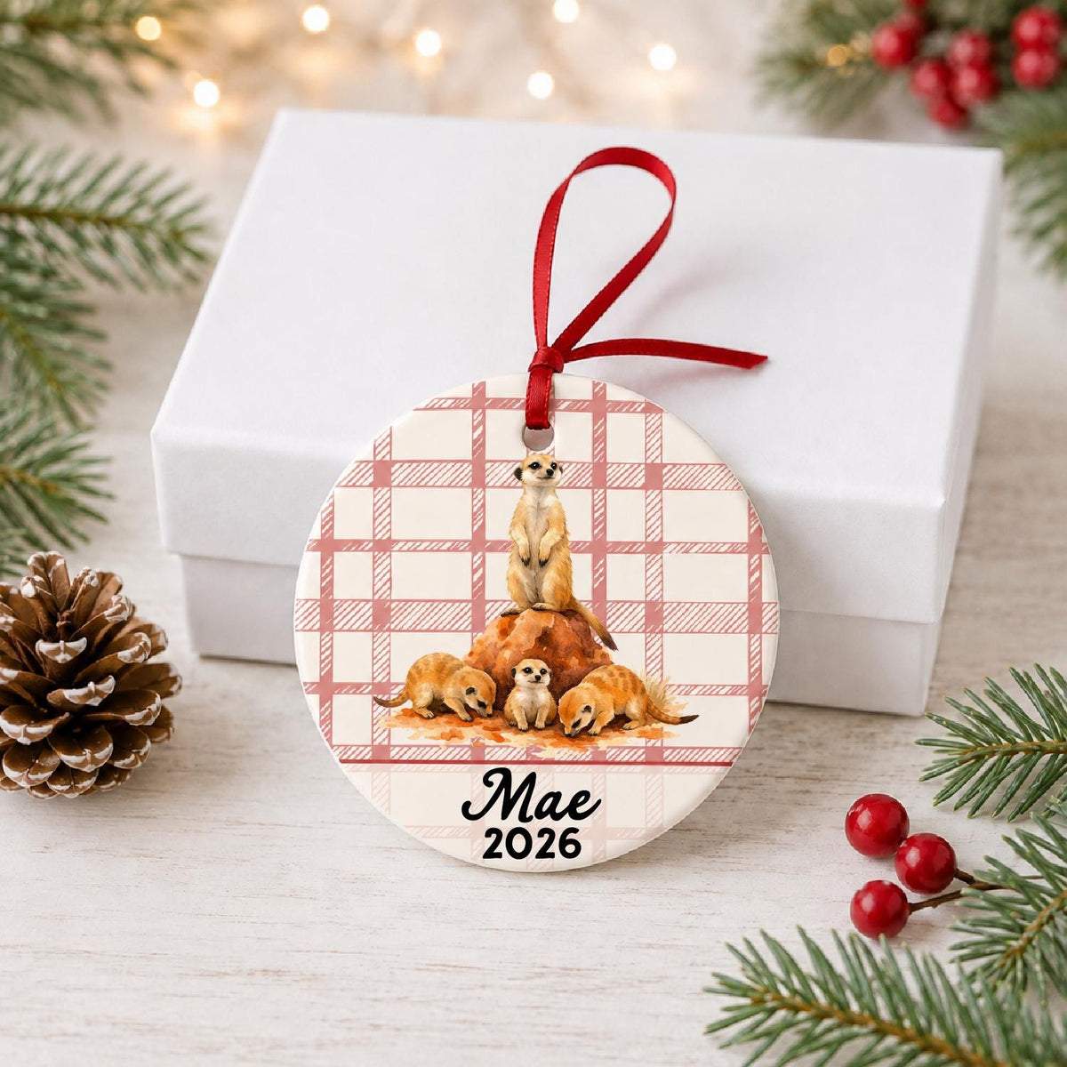 Personalized meerkat ceramic ornament with name and year on red plaid in front of a white gift box with pinecones and red berries