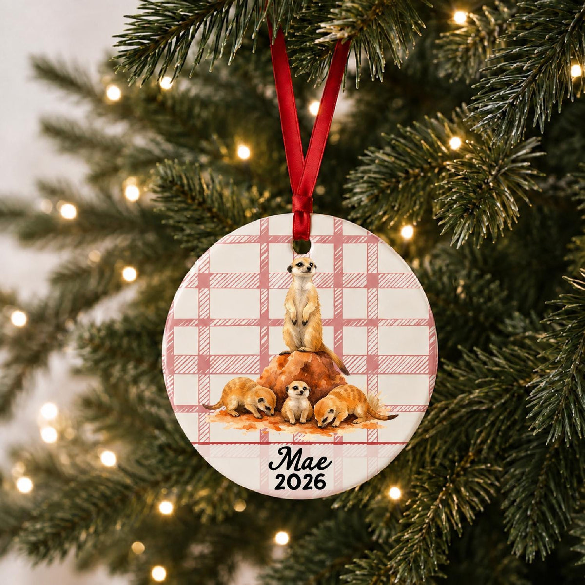 Personalized meerkat ceramic ornament with name and year on red plaid hanging on a lit Christmas tree