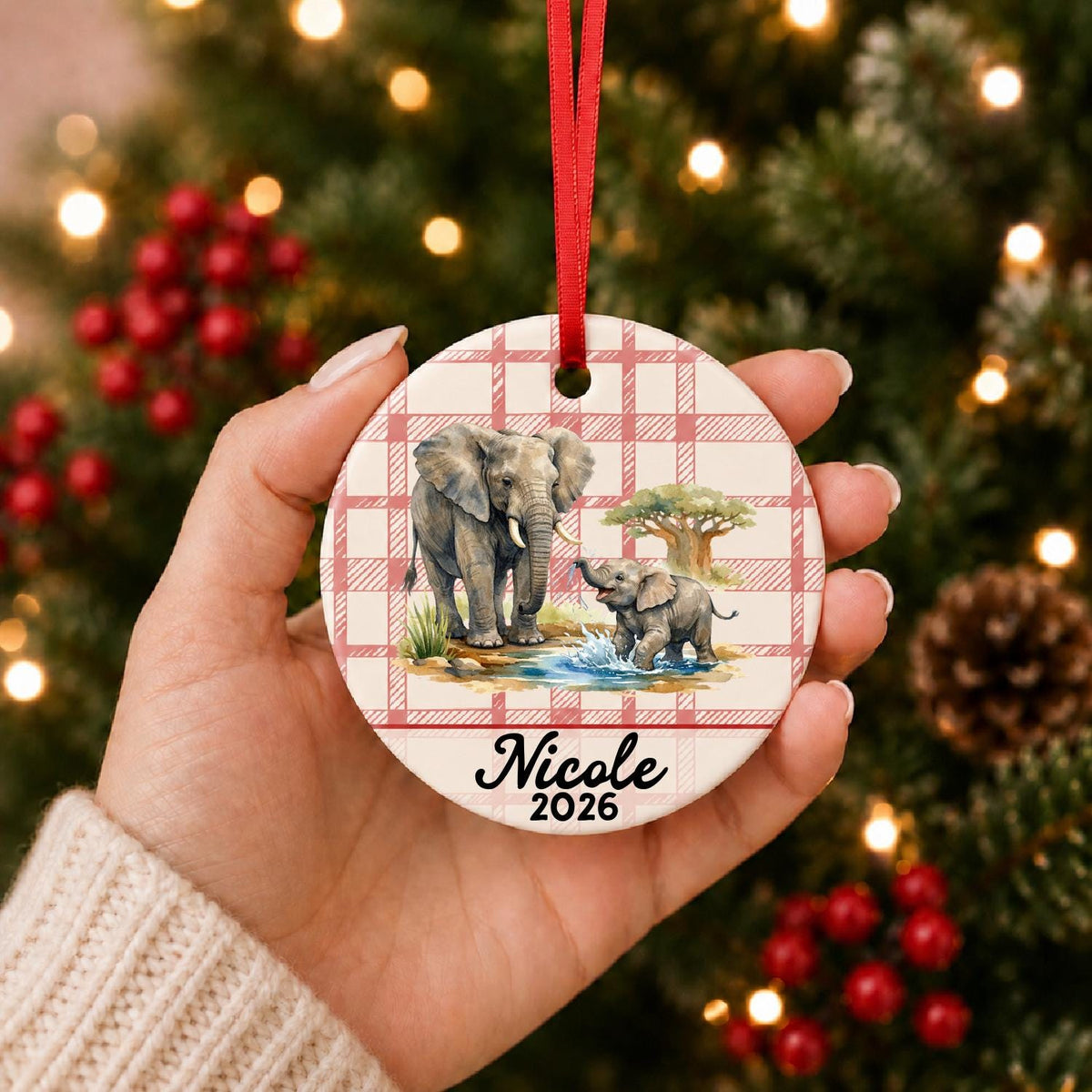 Hand holding the personalized African elephant ceramic ornament with name and year on red plaid in front of a Christmas tree