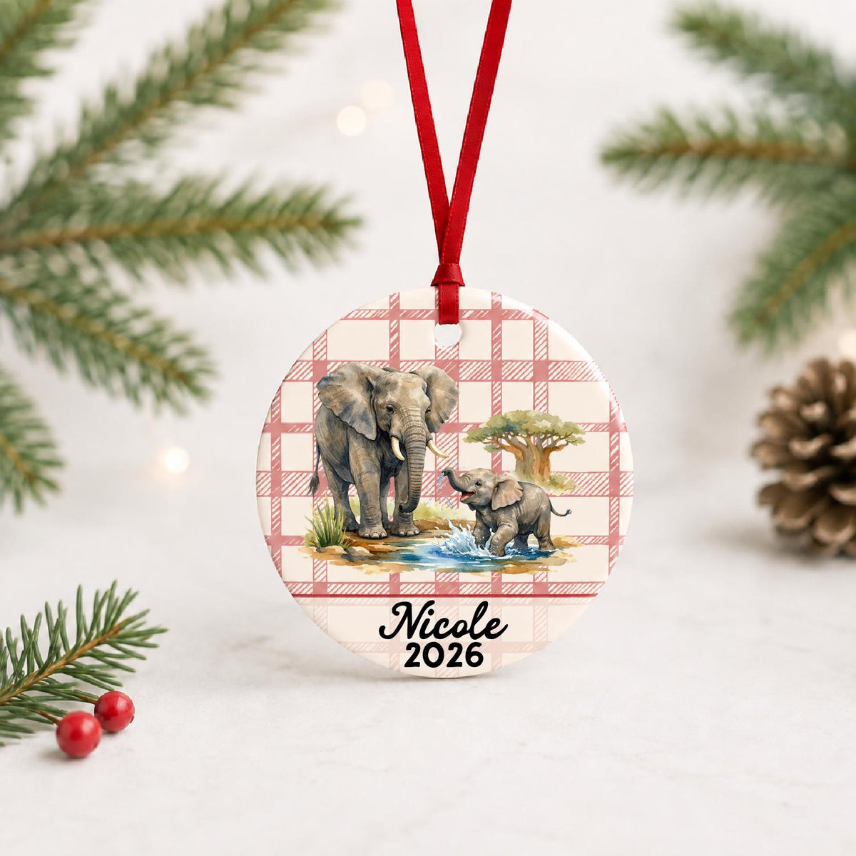 Personalized African elephant ceramic ornament with name and year on red plaid with red ribbon on a white surface with pine branches and pinecones