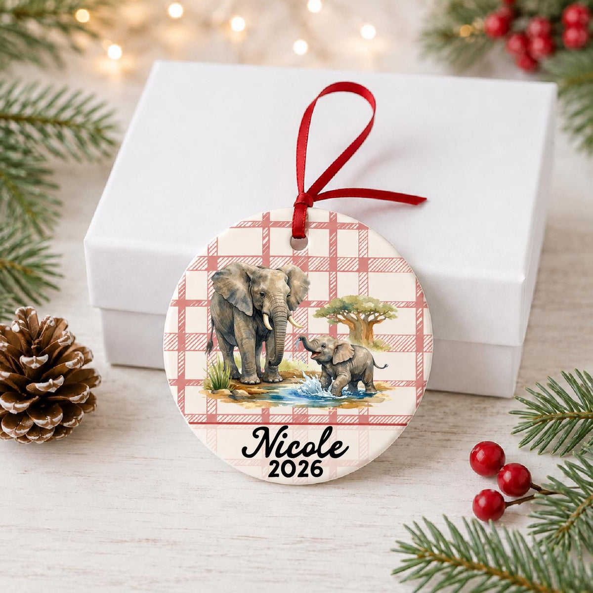 Personalized African elephant ceramic ornament with name and year on red plaid in front of a white gift box with pinecones and red berries
