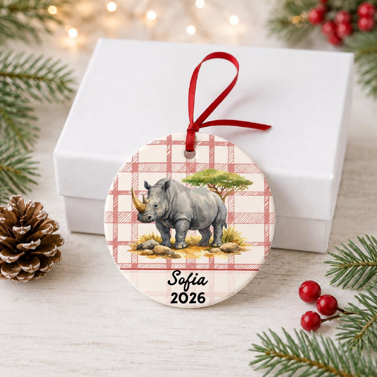 Personalized white rhinoceros ceramic ornament with name and year on red plaid in front of a white gift box with pinecones and red berries