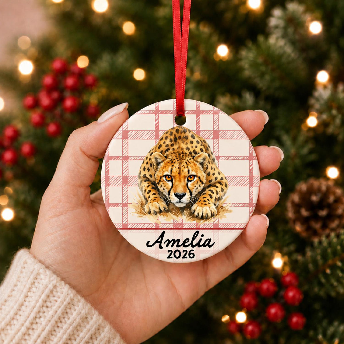Hand holding the personalized cheetah ceramic ornament with name and year on red plaid in front of a Christmas tree
