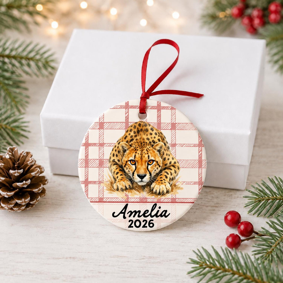 Personalized cheetah ceramic ornament with name and year on red plaid in front of a white gift box with pinecones and red berries
