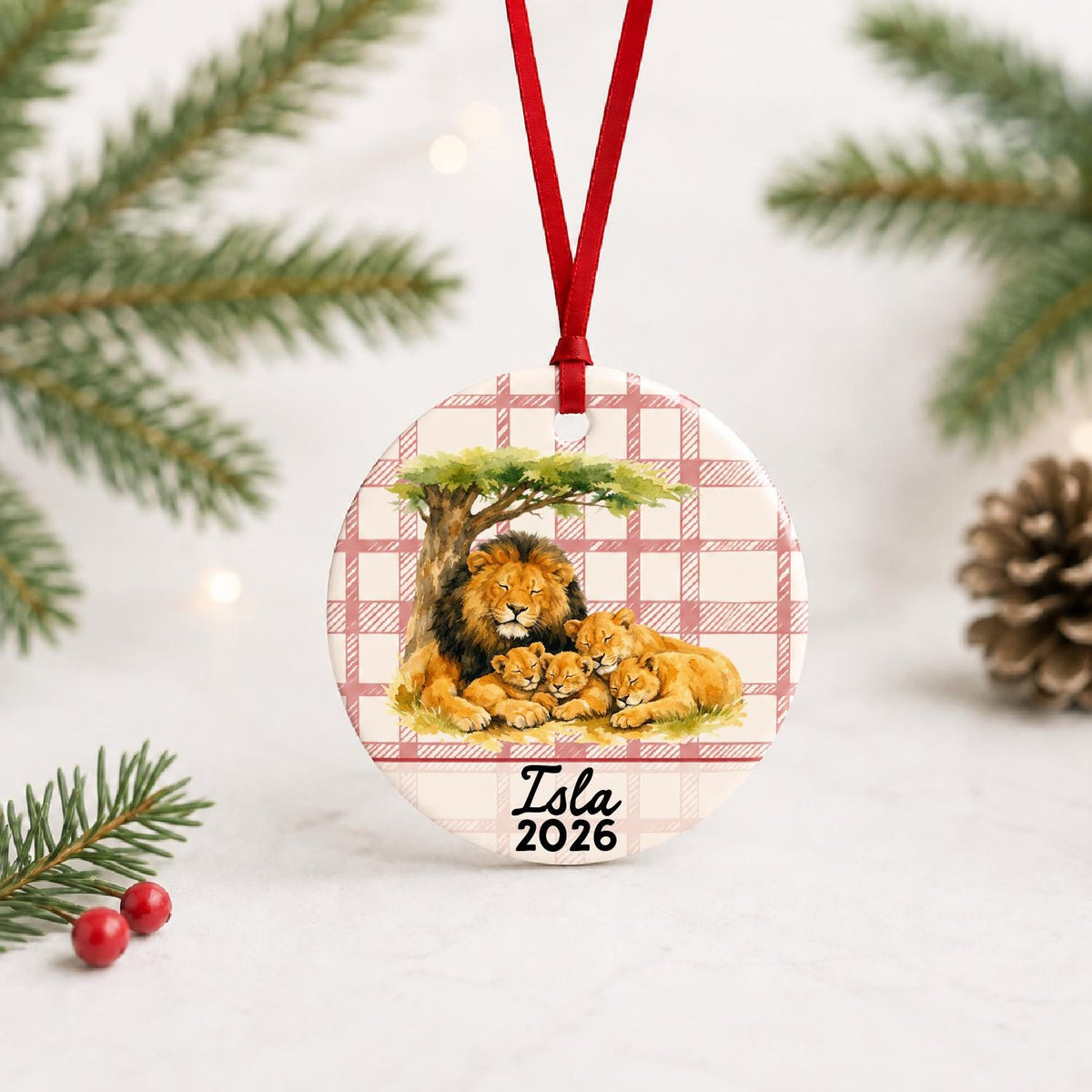 Personalized lion pride ceramic ornament with name and year on red plaid with red ribbon on a white surface with pine branches and pinecones