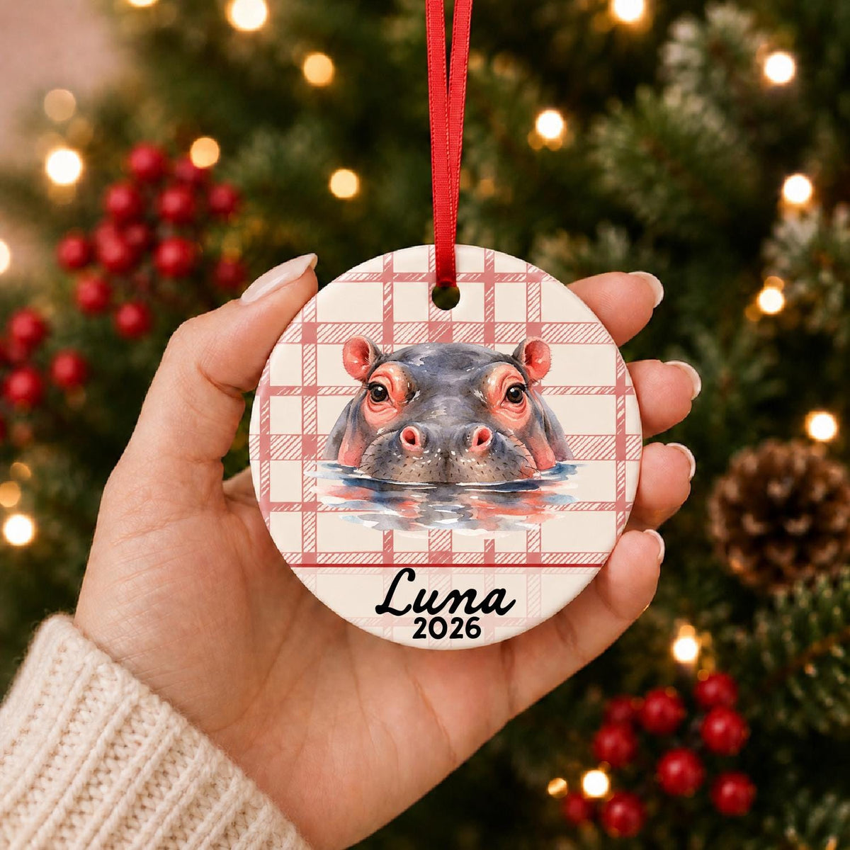 Hand holding the personalized hippopotamus ceramic ornament with name and year on red plaid in front of a Christmas tree