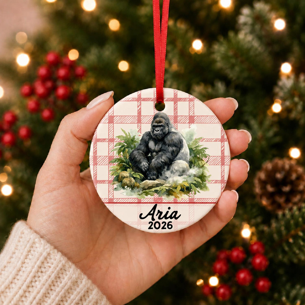Hand holding the personalized silverback gorilla ceramic ornament with name and year on red plaid in front of a Christmas tree