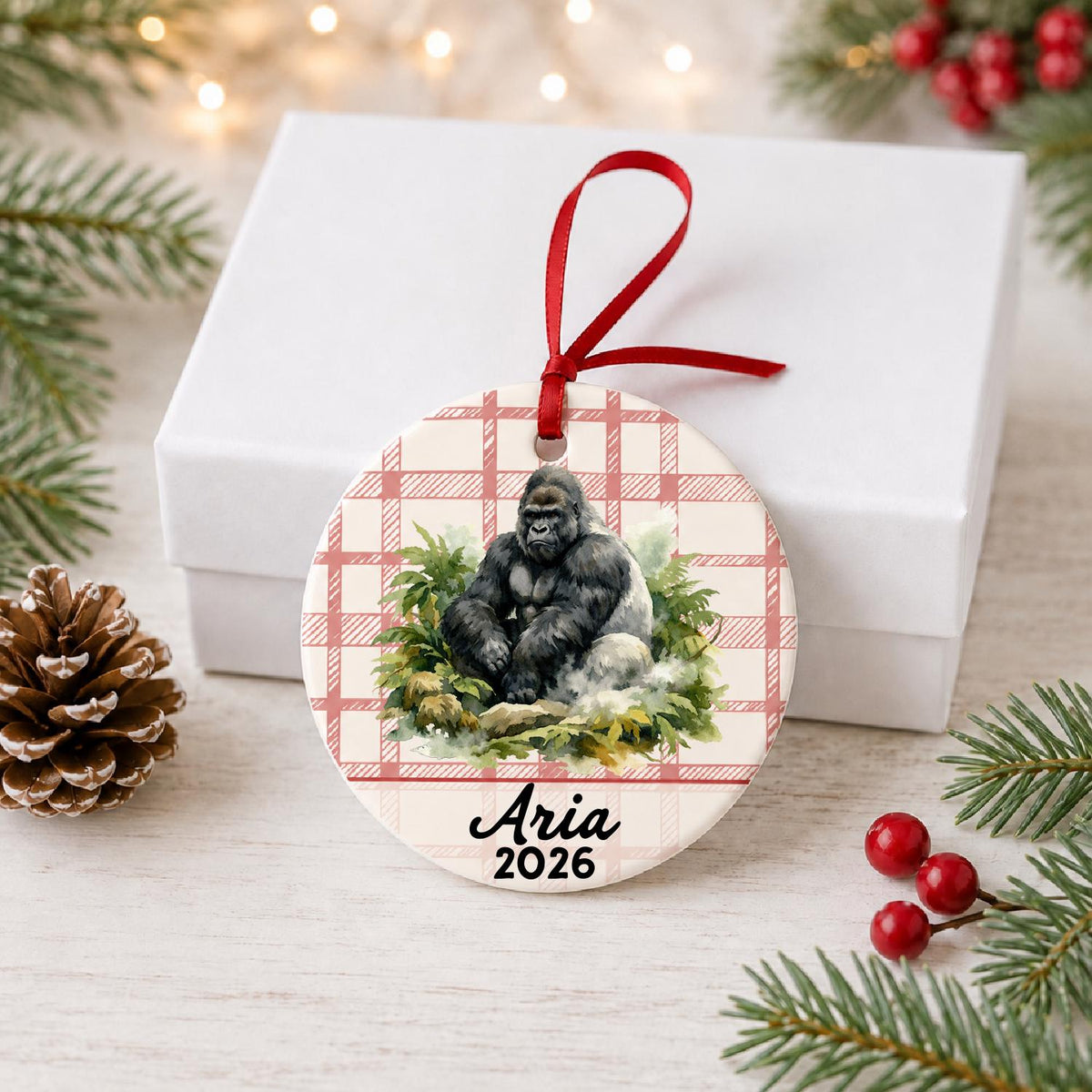 Personalized silverback gorilla ceramic ornament with name and year on red plaid in front of a white gift box with pinecones and red berries
