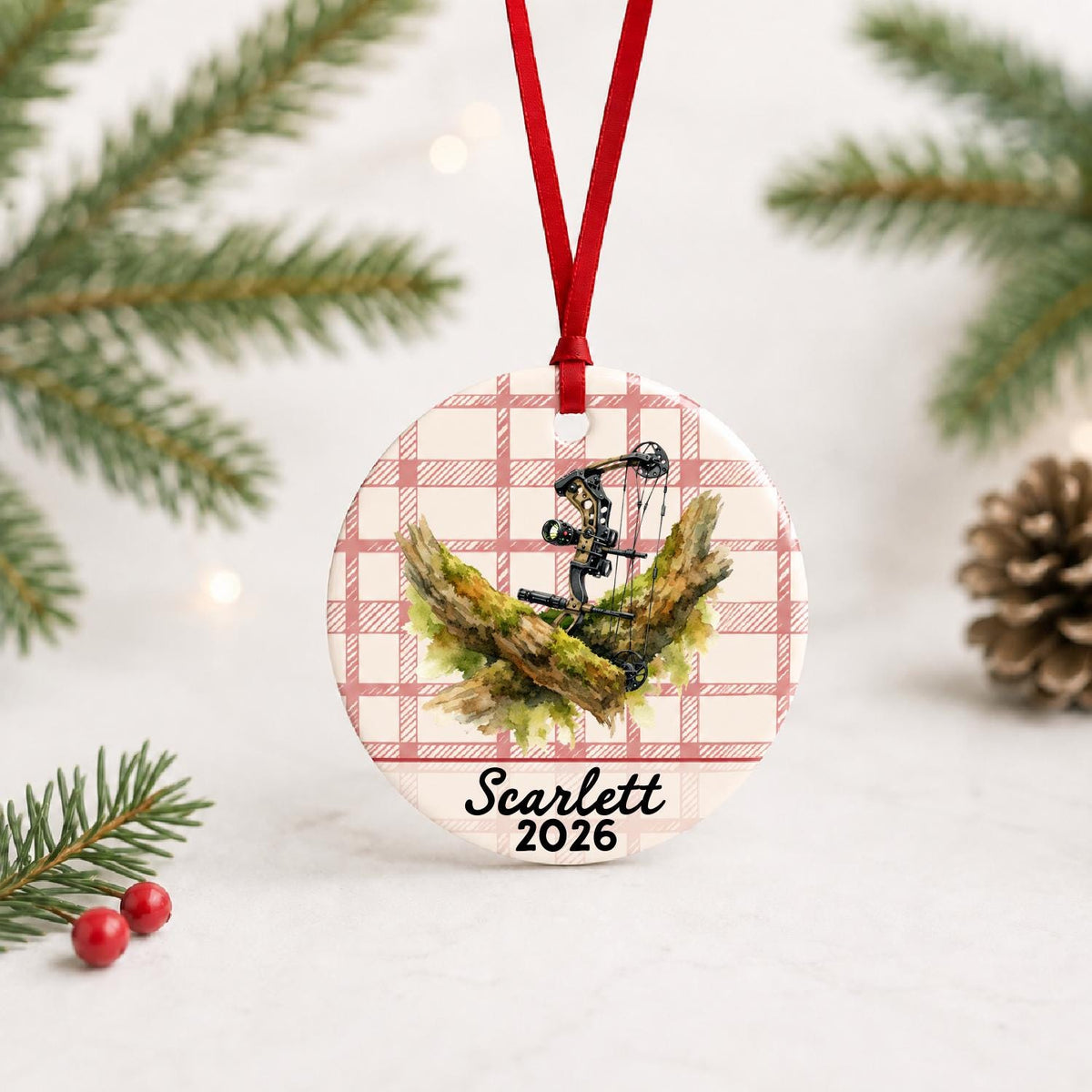 Personalized compound bow ceramic ornament with name and year on red plaid with red ribbon on a white surface with pine branches and pinecones