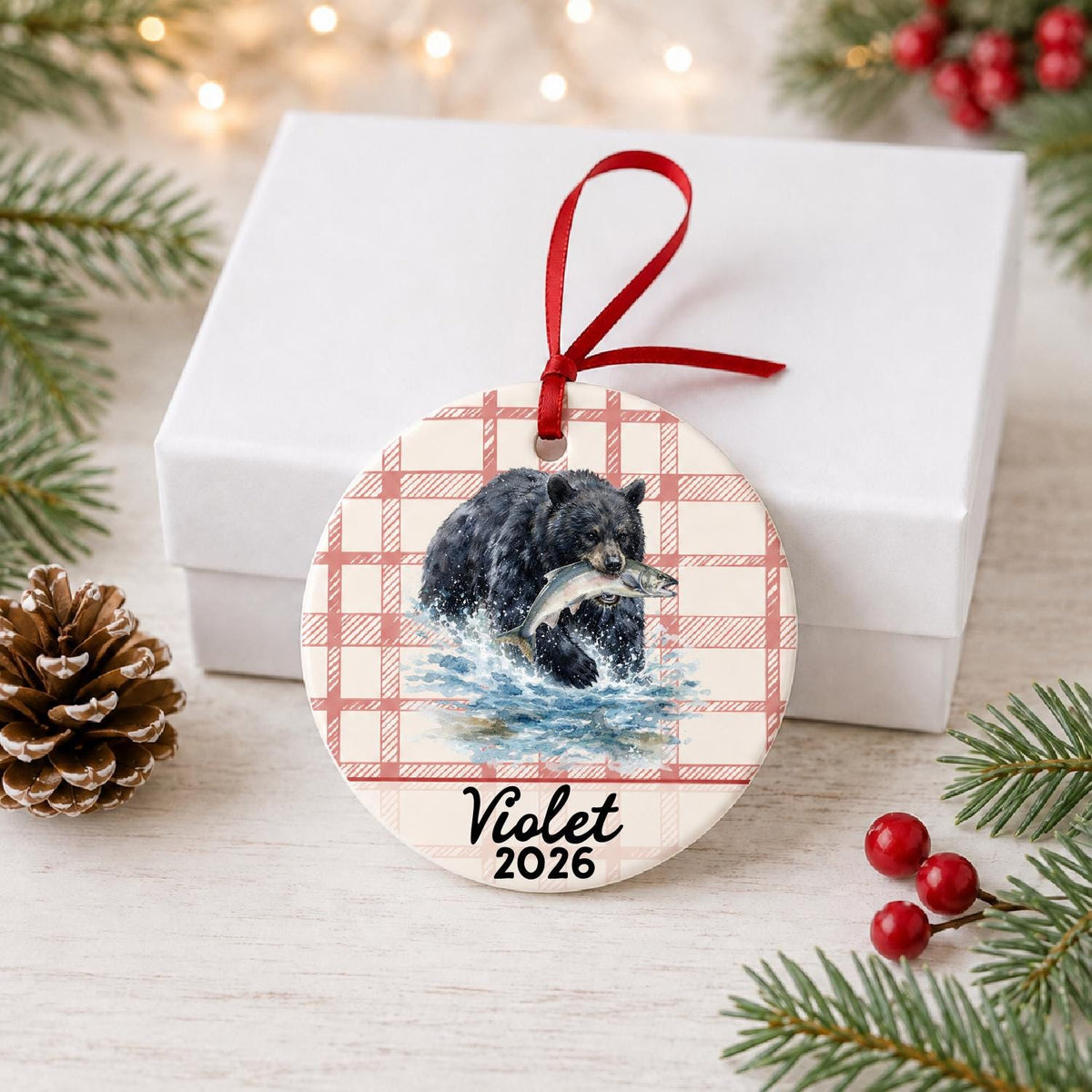 Personalized American black bear ceramic ornament with name and year on red plaid in front of a white gift box with pinecones and red berries