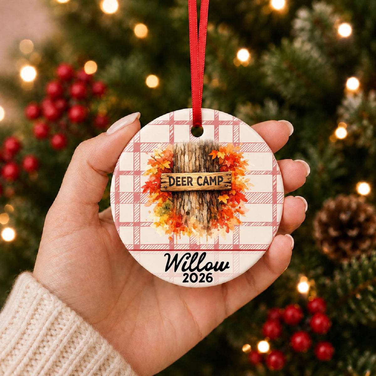 Hand holding the personalized deer camp ceramic ornament with name and year on red plaid in front of a Christmas tree