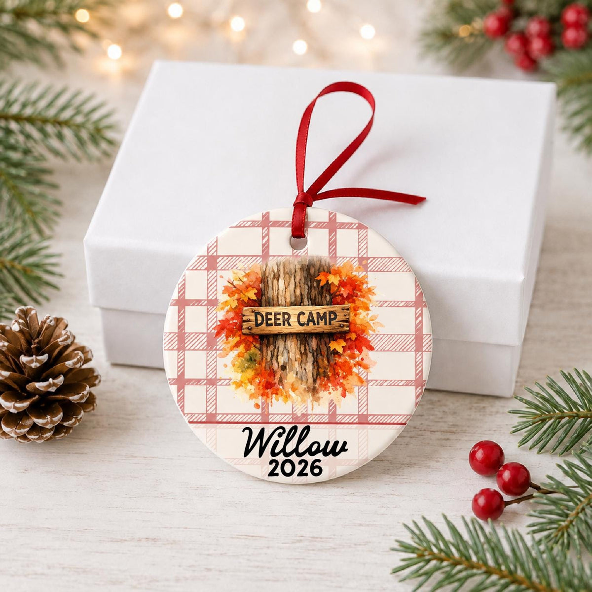 Personalized deer camp ceramic ornament with name and year on red plaid in front of a white gift box with pinecones and red berries
