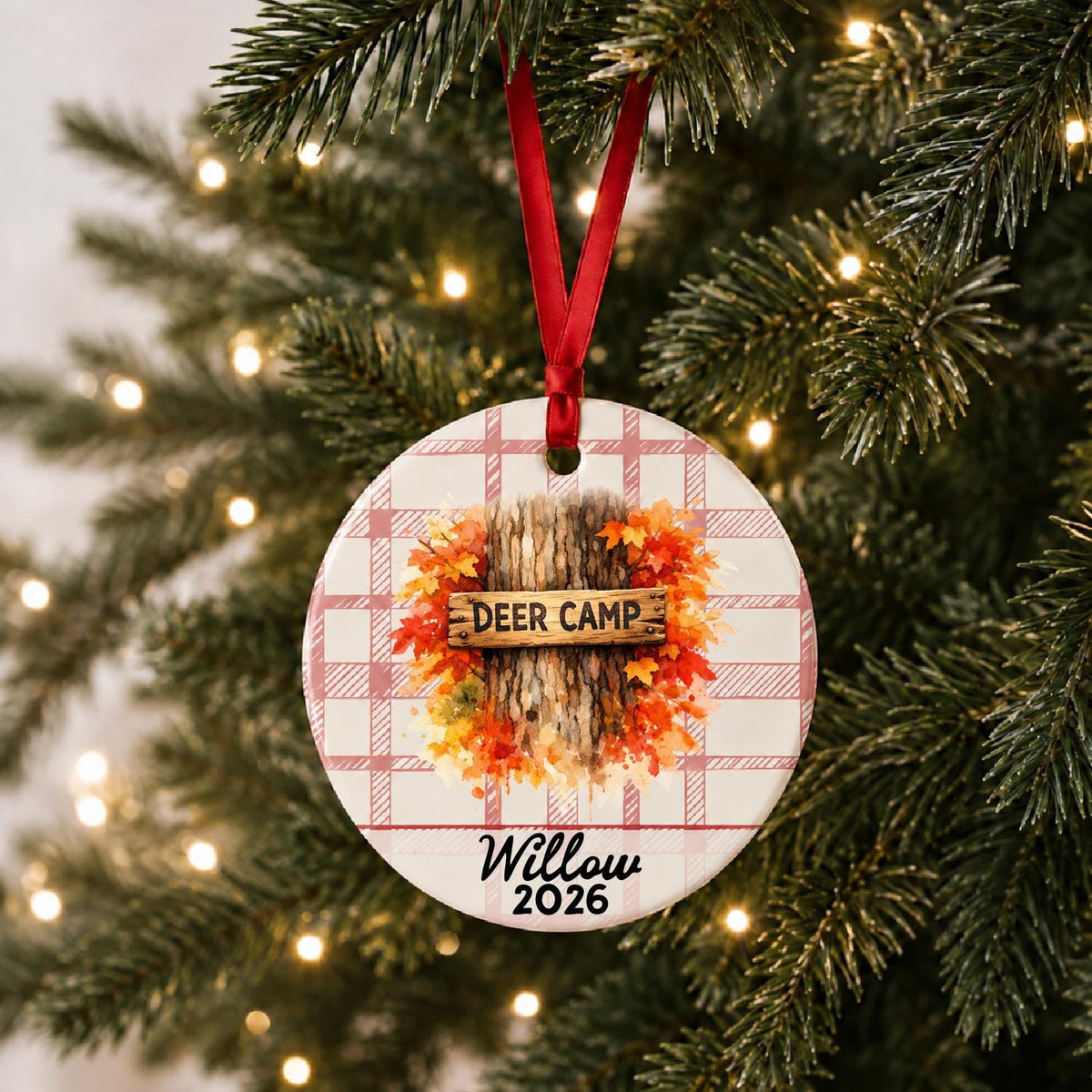 Personalized deer camp ceramic ornament with name and year on red plaid hanging on a lit Christmas tree