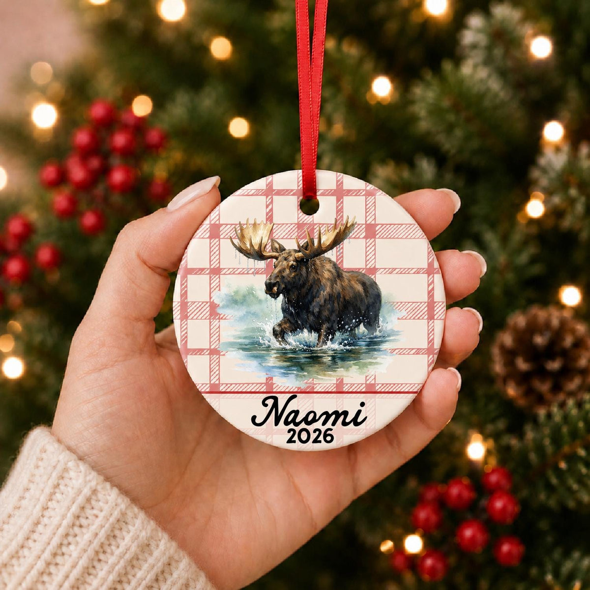 Hand holding the personalized bull moose ceramic ornament with name and year on red plaid in front of a Christmas tree