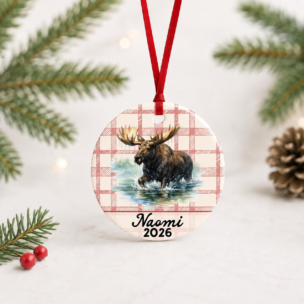 Personalized bull moose ceramic ornament with name and year on red plaid with red ribbon on a white surface with pine branches and pinecones