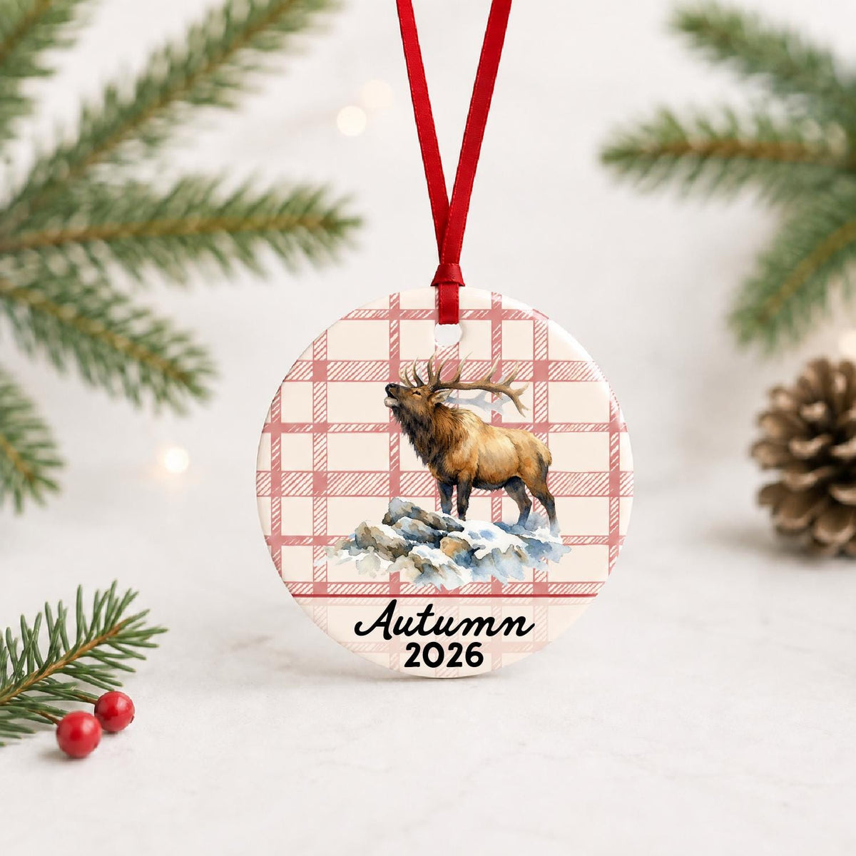 Personalized bull elk ceramic ornament with name and year on red plaid with red ribbon on a white surface with pine branches and pinecones
