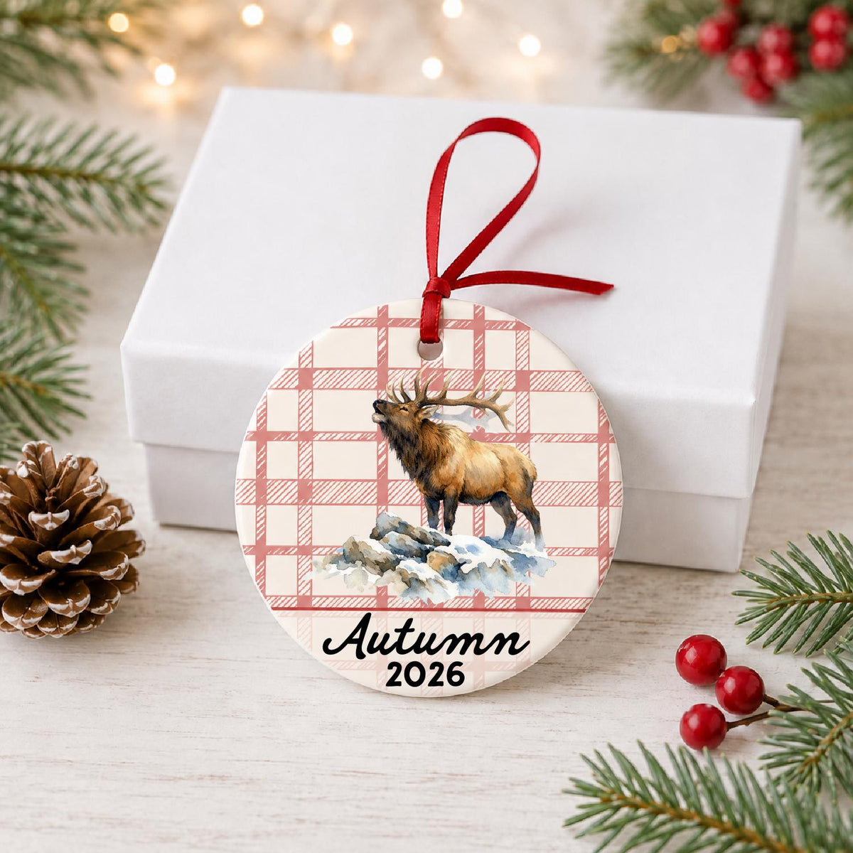 Personalized bull elk ceramic ornament with name and year on red plaid in front of a white gift box with pinecones and red berries