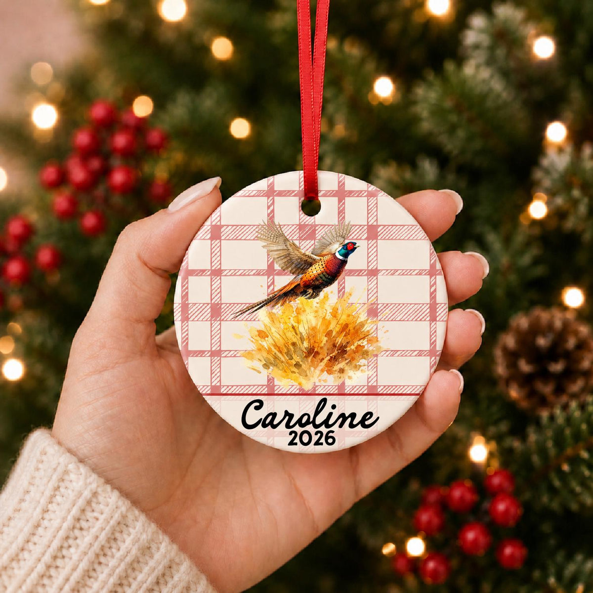Hand holding the personalized ring-necked pheasant ceramic ornament with name and year on red plaid in front of a Christmas tree