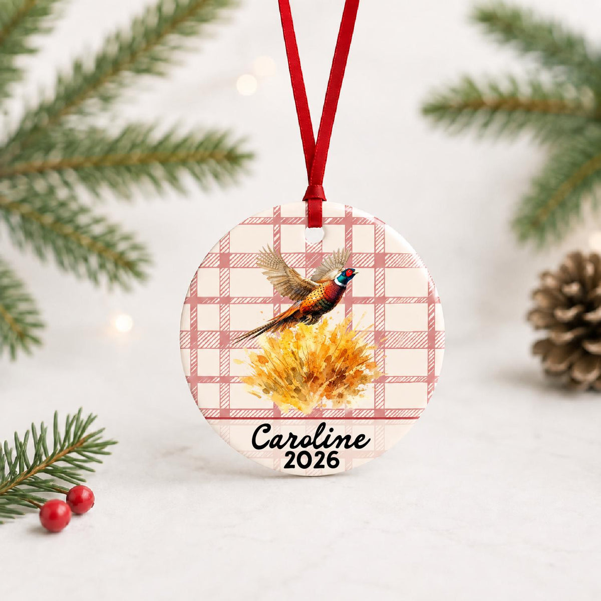 Personalized ring-necked pheasant ceramic ornament with name and year on red plaid with red ribbon on a white surface with pine branches and pinecones
