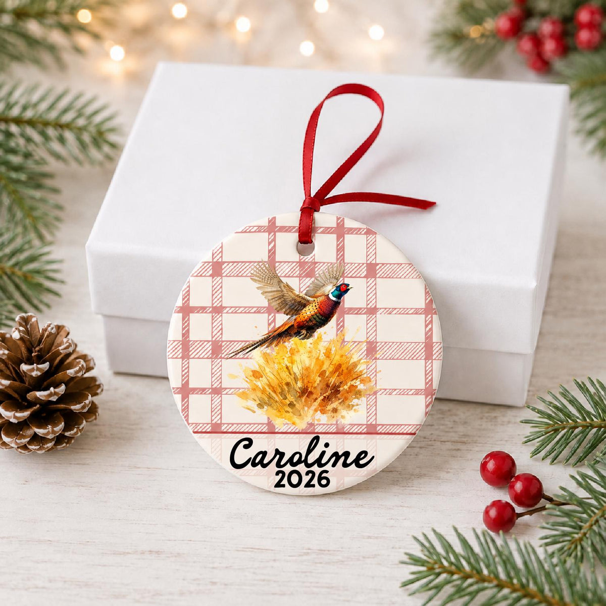 Personalized ring-necked pheasant ceramic ornament with name and year on red plaid in front of a white gift box with pinecones and red berries