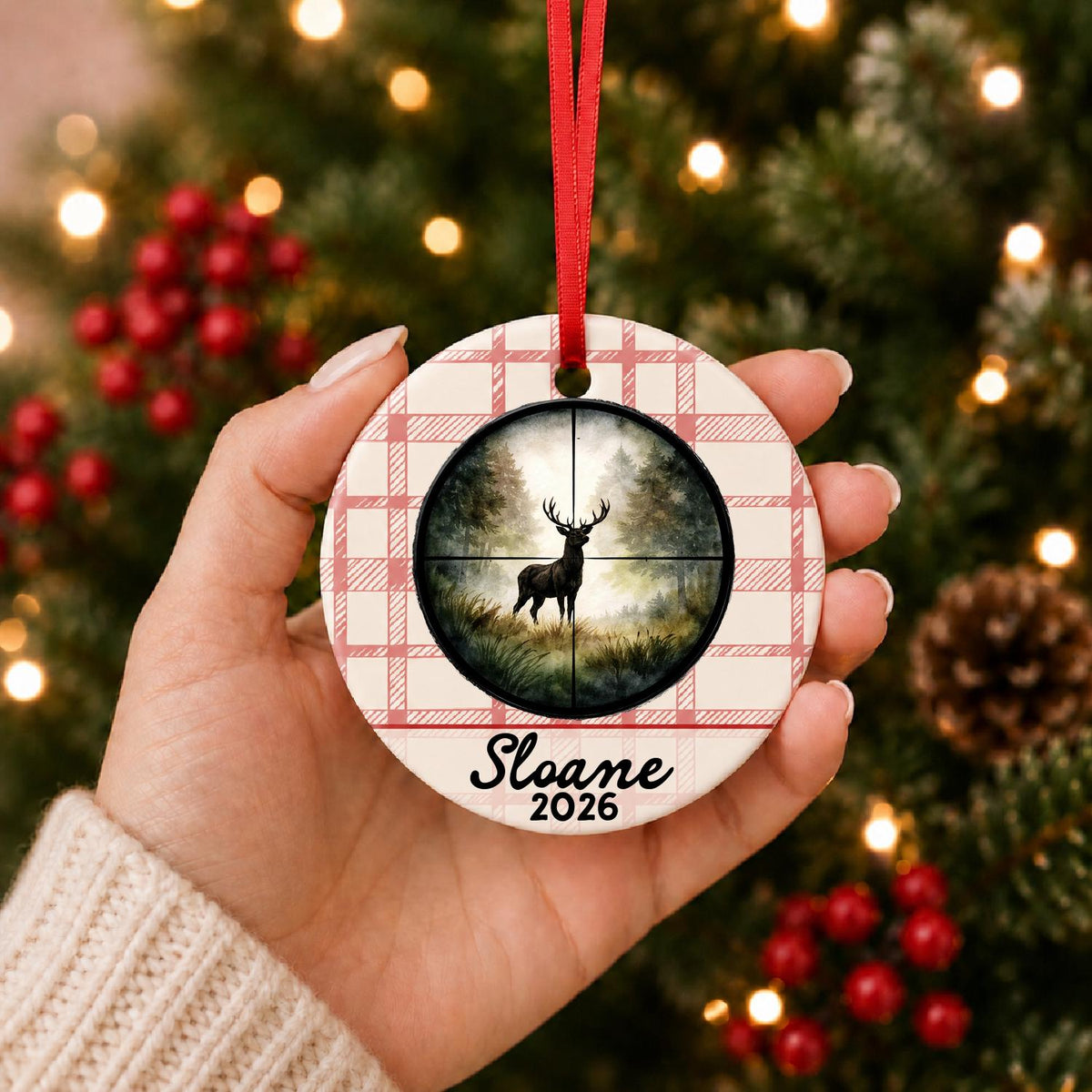 Hand holding the personalized deer hunting ceramic ornament with name and year on red plaid in front of a Christmas tree
