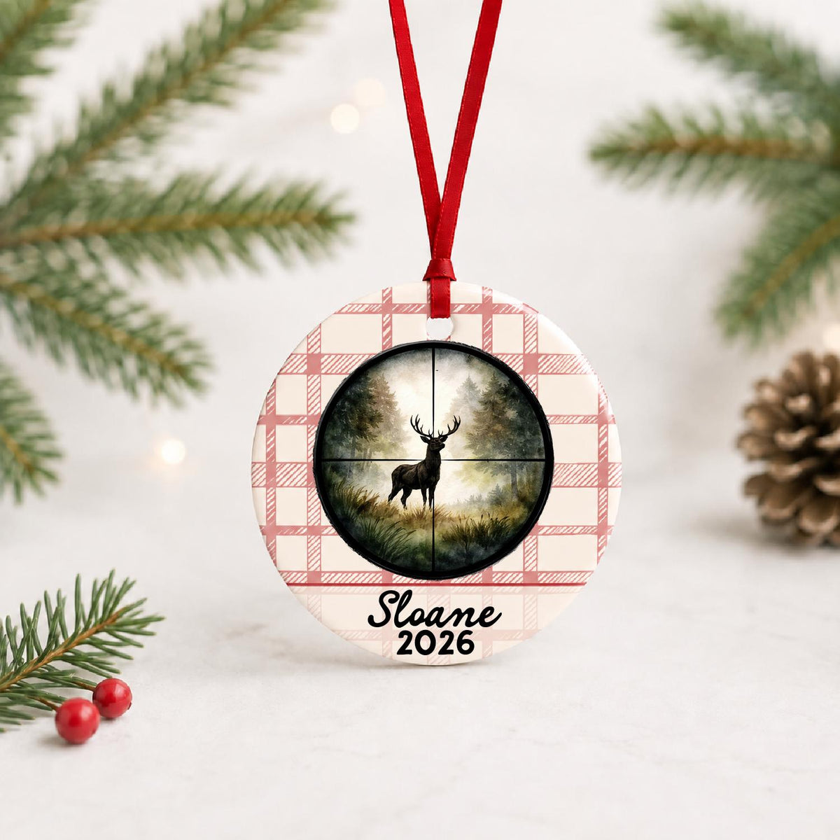 Personalized deer hunting ceramic ornament with name and year on red plaid with red ribbon on a white surface with pine branches and pinecones