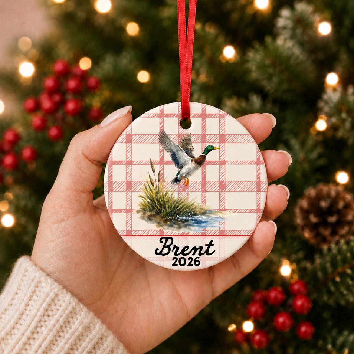 Hand holding the personalized mallard duck ceramic ornament with name and year on red plaid in front of a Christmas tree