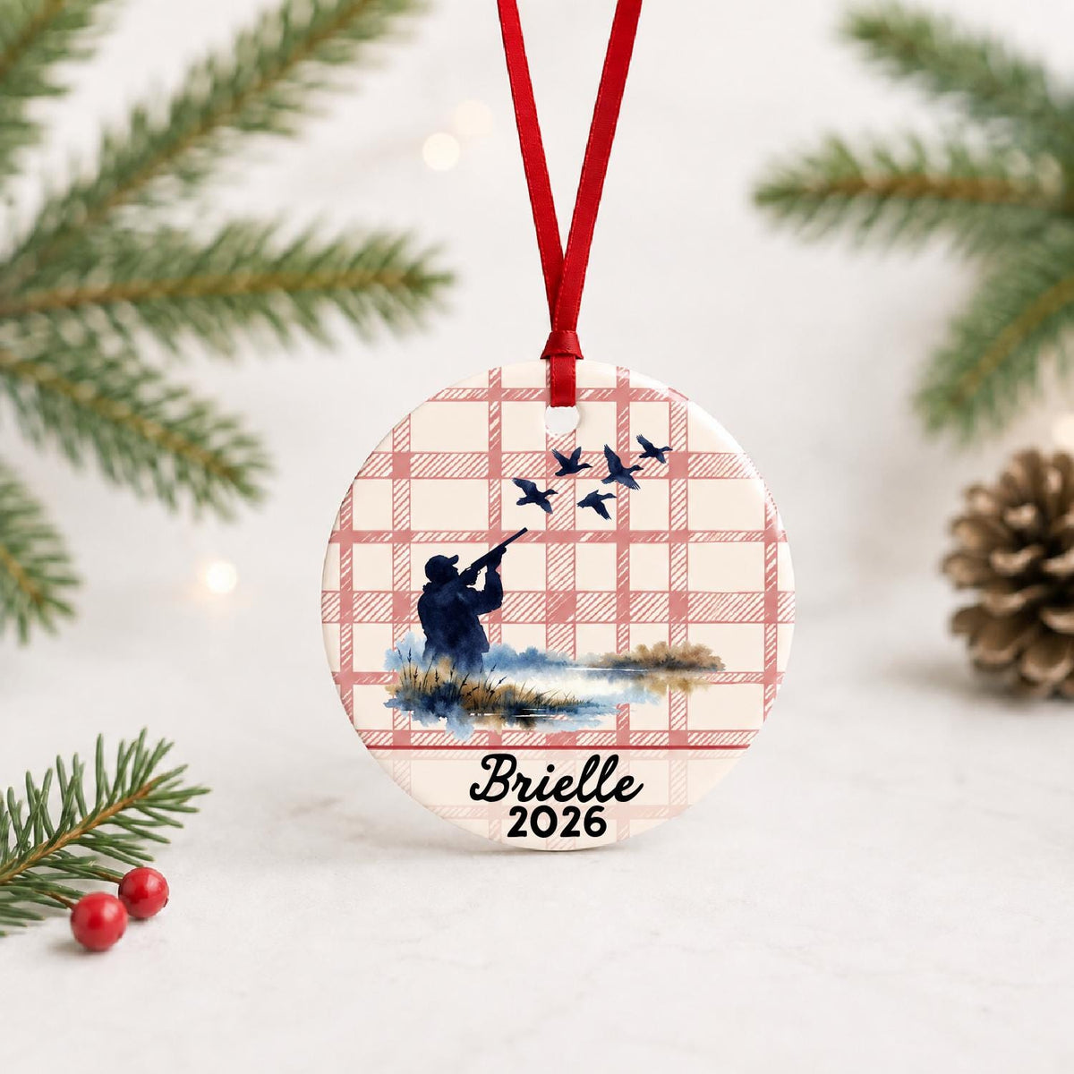 Personalized duck hunting ceramic ornament with name and year on red plaid with red ribbon on a white surface with pine branches and pinecones
