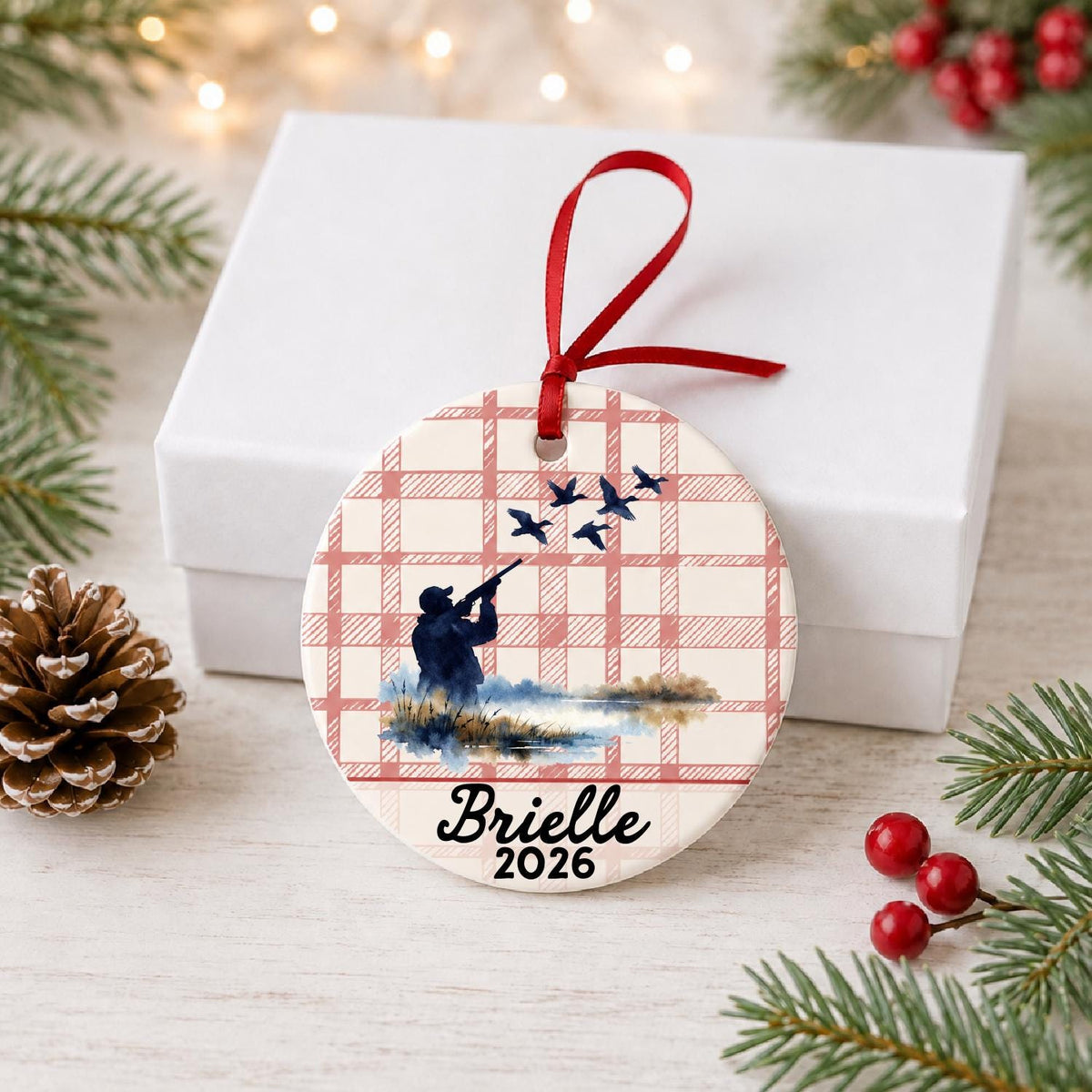 Personalized duck hunting ceramic ornament with name and year on red plaid in front of a white gift box with pinecones and red berries