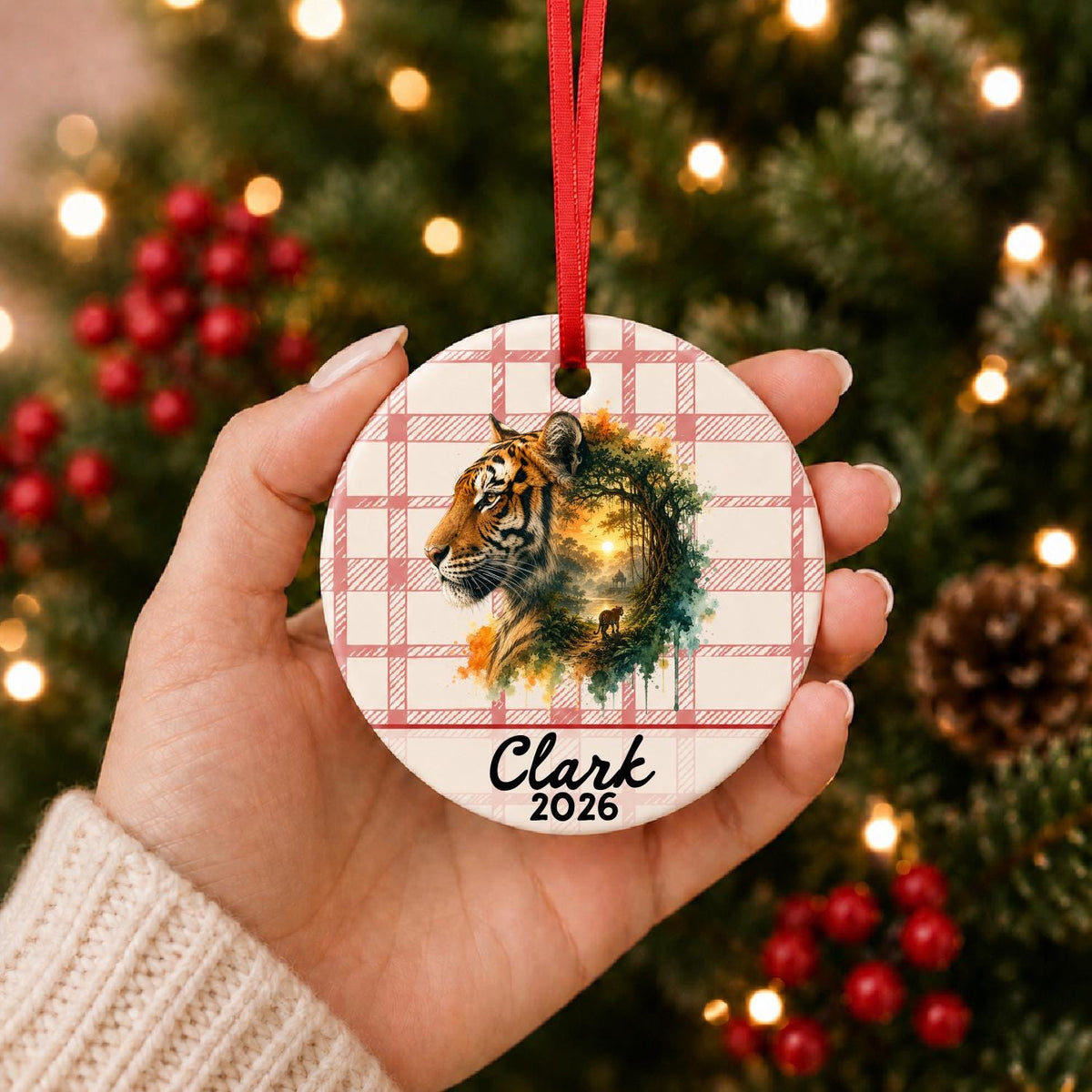 Hand holding the personalized tiger ceramic ornament with name and year on red plaid in front of a Christmas tree