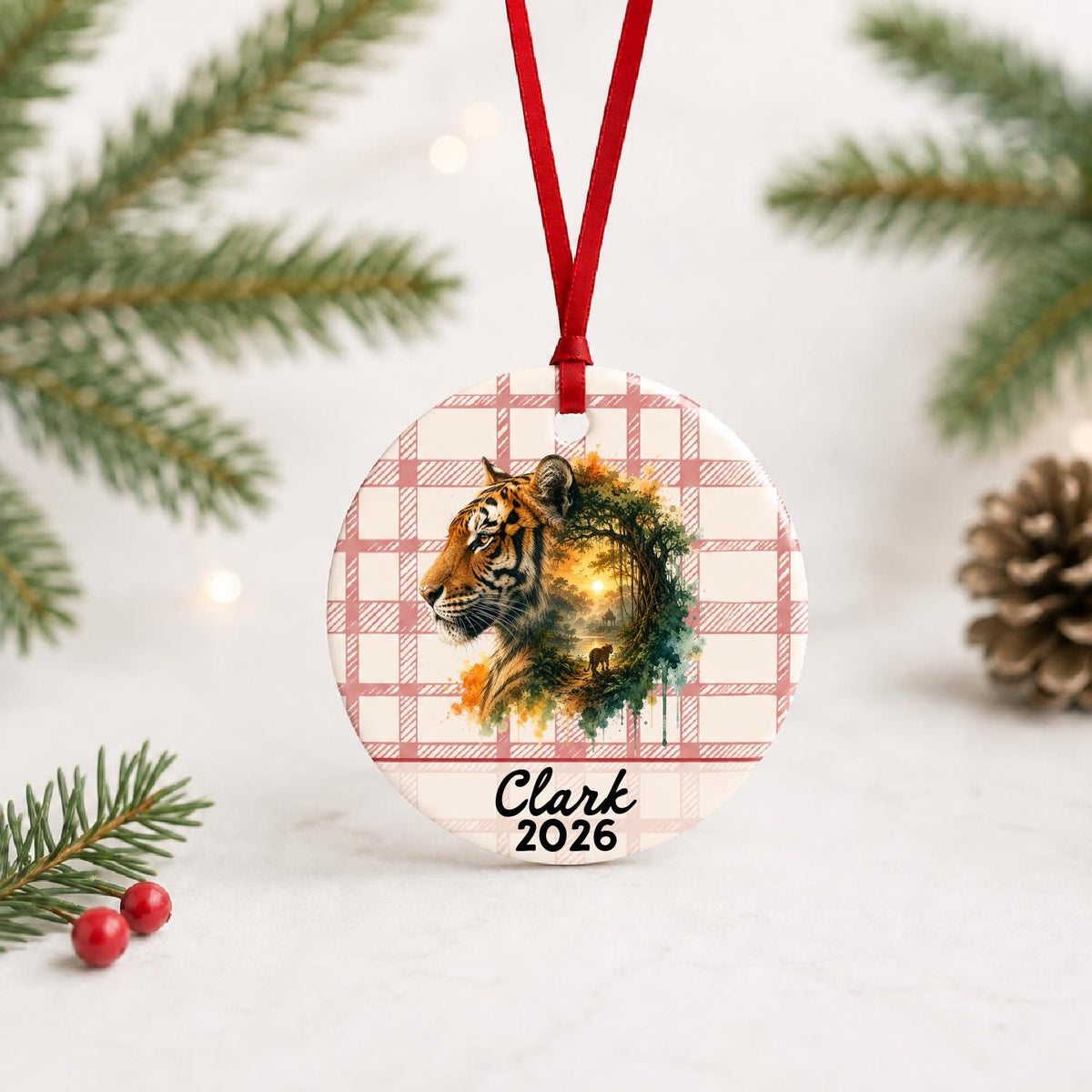 Personalized tiger ceramic ornament with name and year on red plaid with red ribbon on a white surface with pine branches and pinecones
