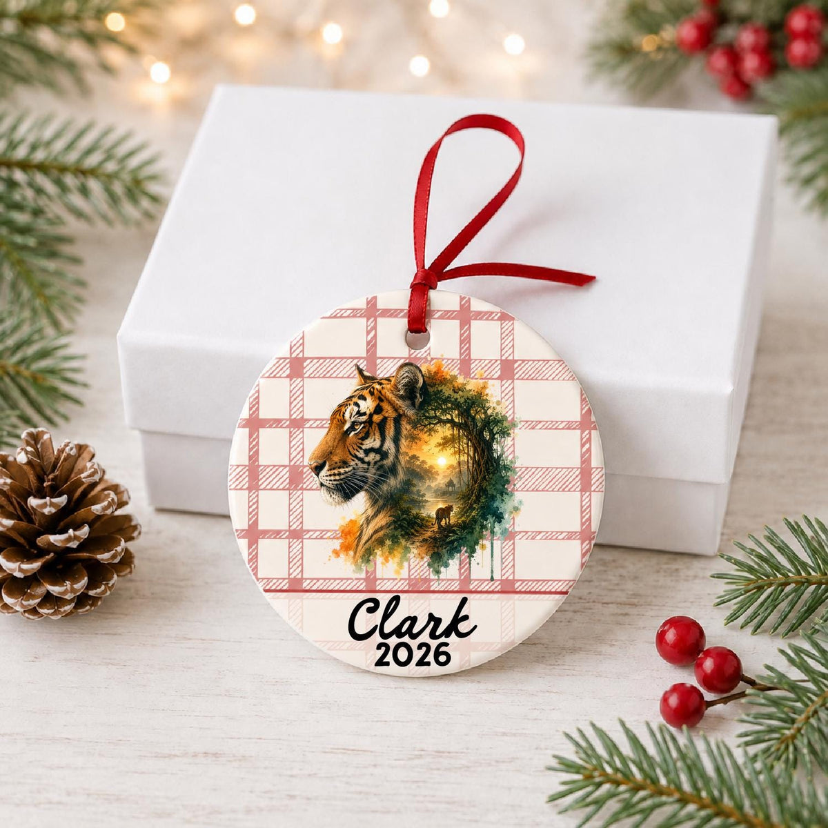 Personalized tiger ceramic ornament with name and year on red plaid in front of a white gift box with pinecones and red berries