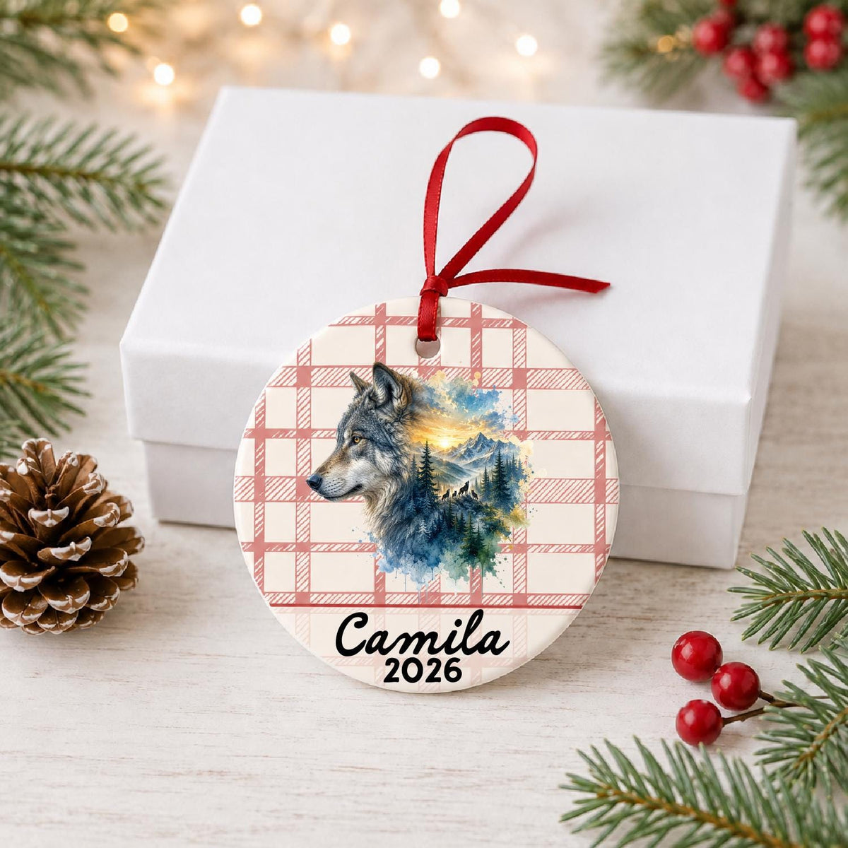 Personalized grey wolf ceramic ornament with name and year on red plaid in front of a white gift box with pinecones and red berries