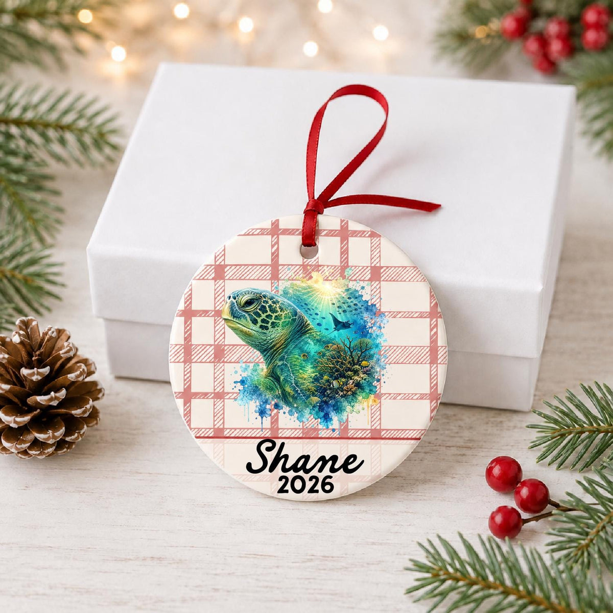 Personalized green sea turtle ceramic ornament with name and year on red plaid in front of a white gift box with pinecones and red berries