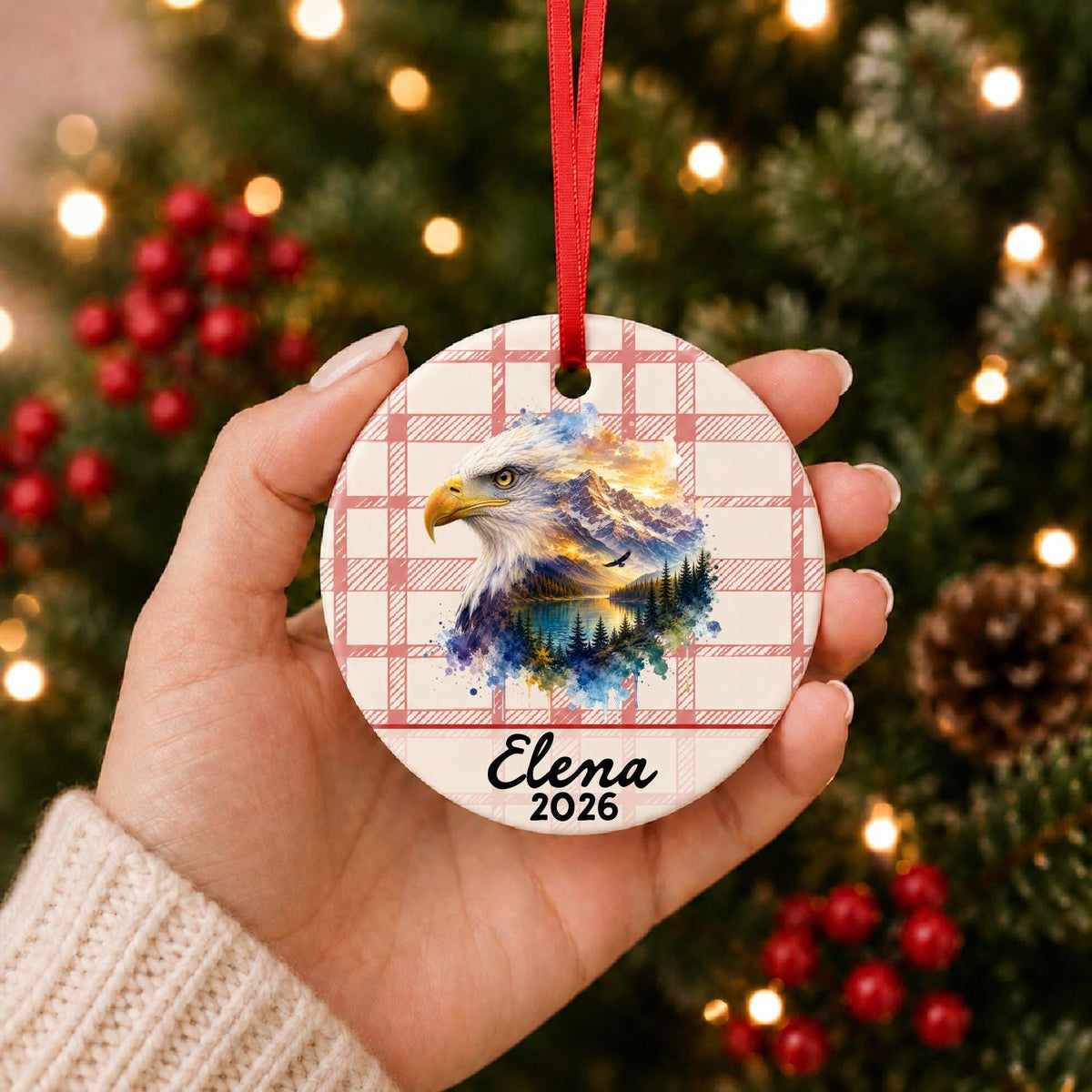 Hand holding the personalized bald eagle ceramic ornament with name and year on red plaid in front of a Christmas tree