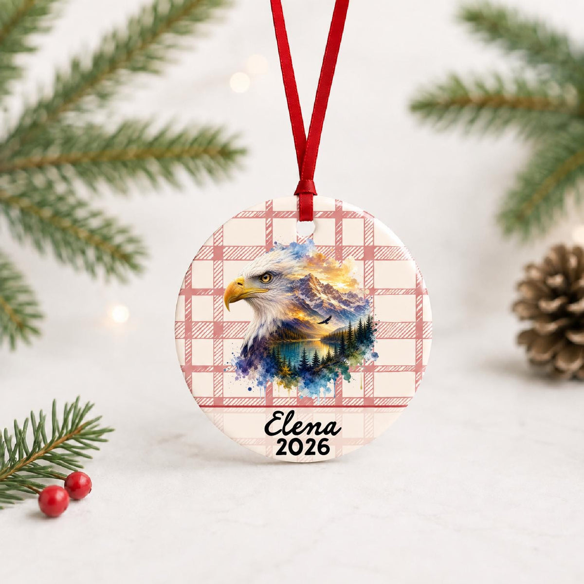 Personalized bald eagle ceramic ornament with name and year on red plaid with red ribbon on a white surface with pine branches and pinecones