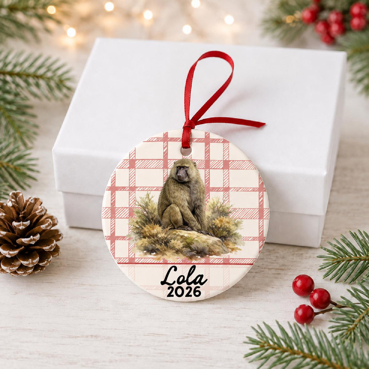 Personalized baboon ceramic ornament with name and year on red plaid in front of a white gift box with pinecones and red berries