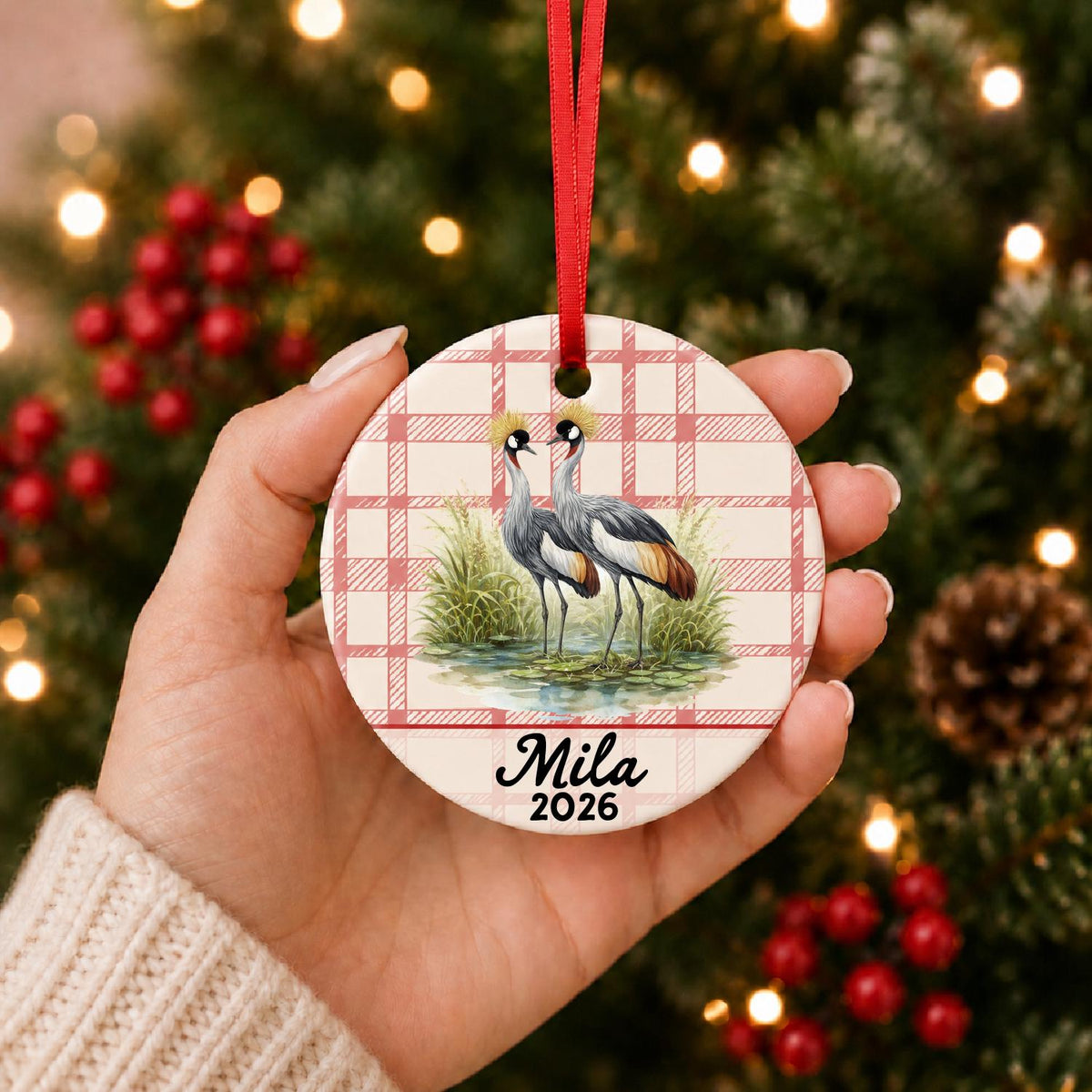 Hand holding the personalized grey crowned crane ceramic ornament with name and year on red plaid in front of a Christmas tree