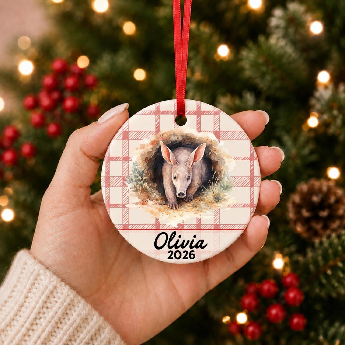 Hand holding the personalized aardvark ceramic ornament with name and year on red plaid in front of a Christmas tree
