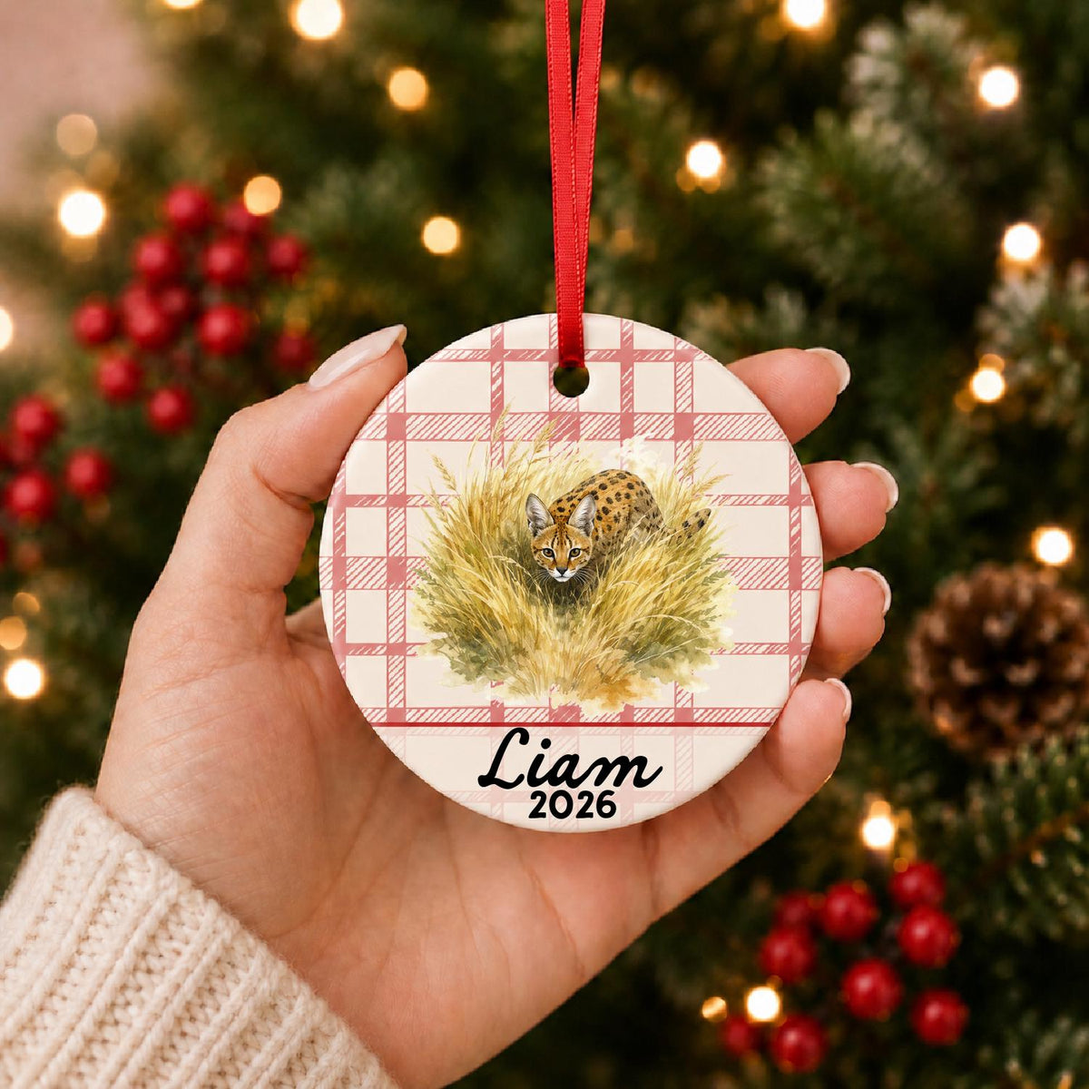 Hand holding the personalized serval ceramic ornament with name and year on red plaid in front of a Christmas tree