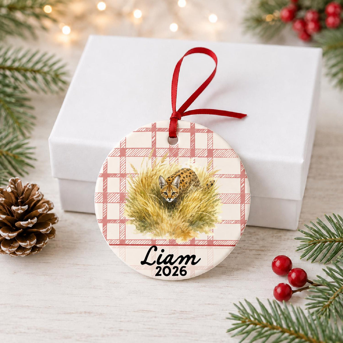 Personalized serval ceramic ornament with name and year on red plaid in front of a white gift box with pinecones and red berries