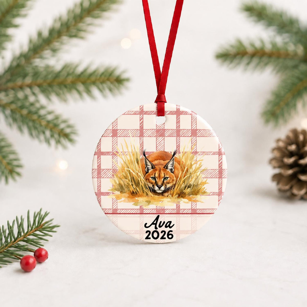 Personalized caracal ceramic ornament with name and year on red plaid with red ribbon on a white surface with pine branches and pinecones