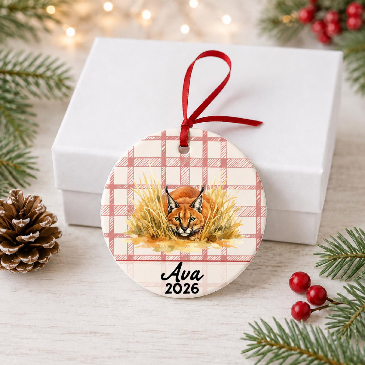 Personalized caracal ceramic ornament with name and year on red plaid in front of a white gift box with pinecones and red berries