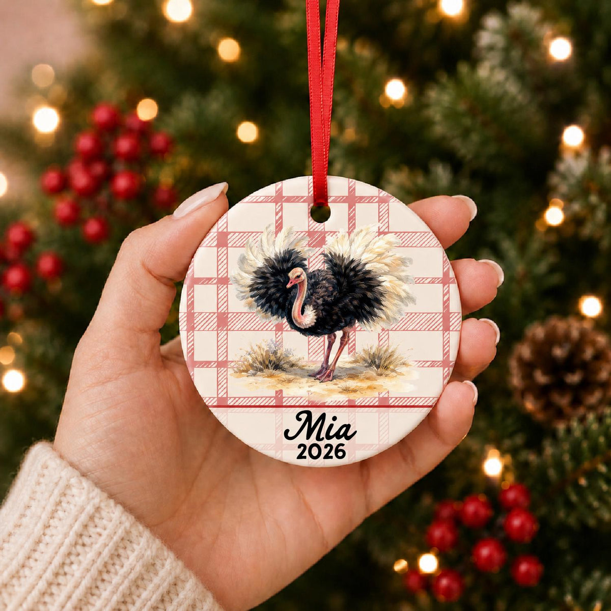 Hand holding the personalized ostrich ceramic ornament with name and year on red plaid in front of a Christmas tree