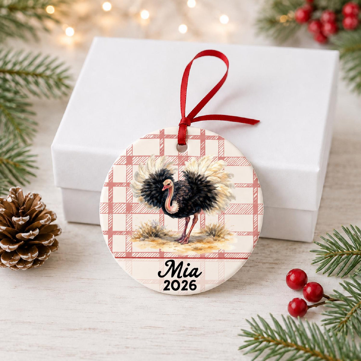 Personalized ostrich ceramic ornament with name and year on red plaid in front of a white gift box with pinecones and red berries