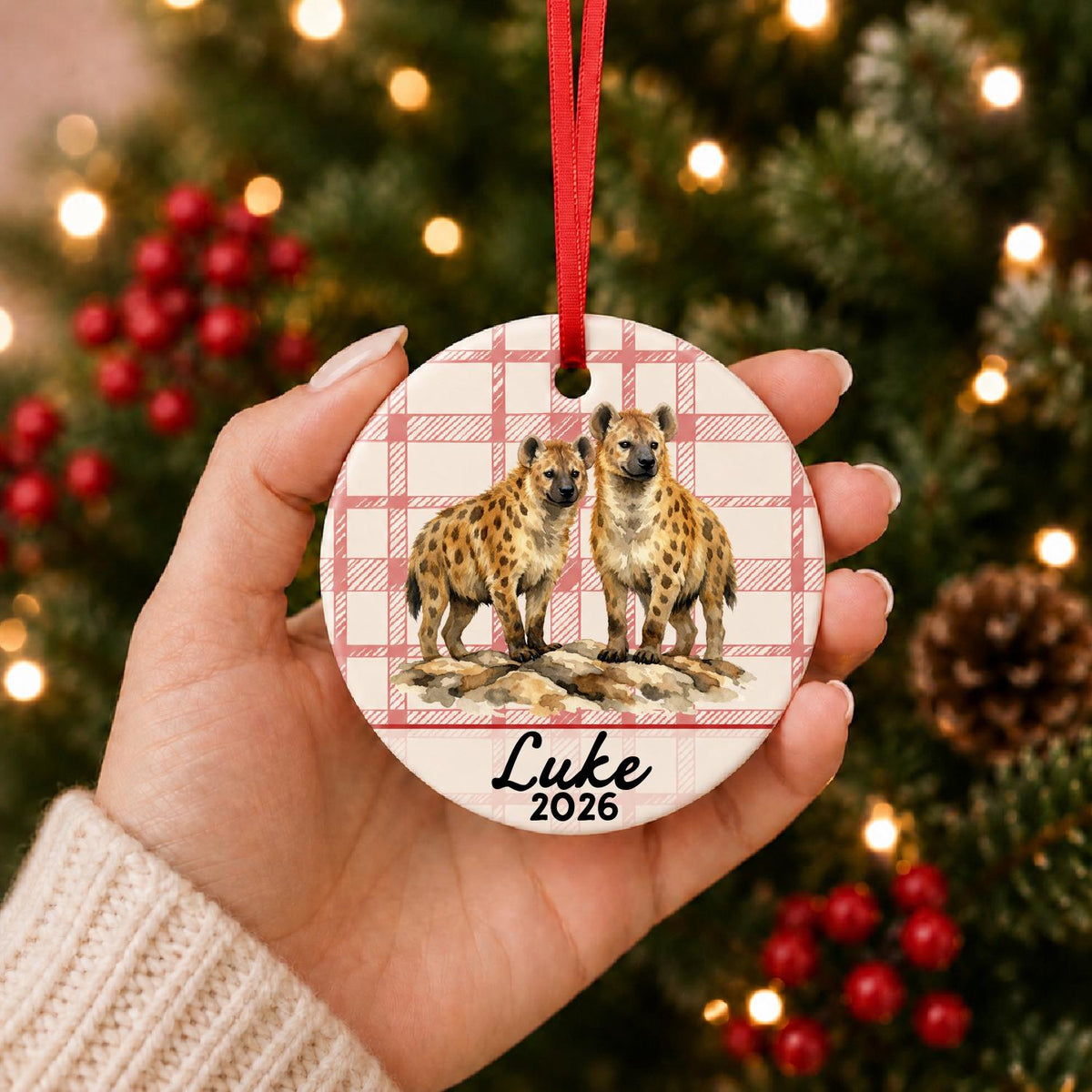 Hand holding the personalized spotted hyena ceramic ornament with name and year on red plaid in front of a Christmas tree